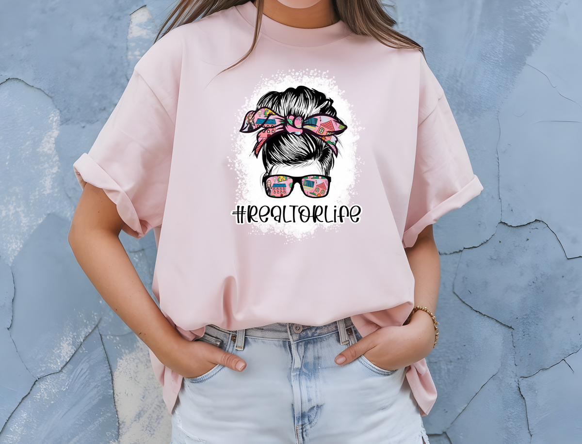 Realtor Inspired Woman’s T-Shirt featuring the message “Realtor Life” with graphic of woman’s face wearing glasses and matching bow and a messy bun”
